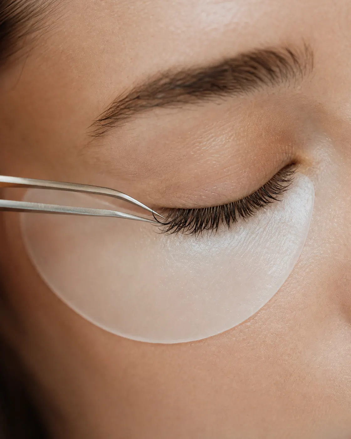 A lash technician applying an individual extension with fine tweezers to a closed eye.